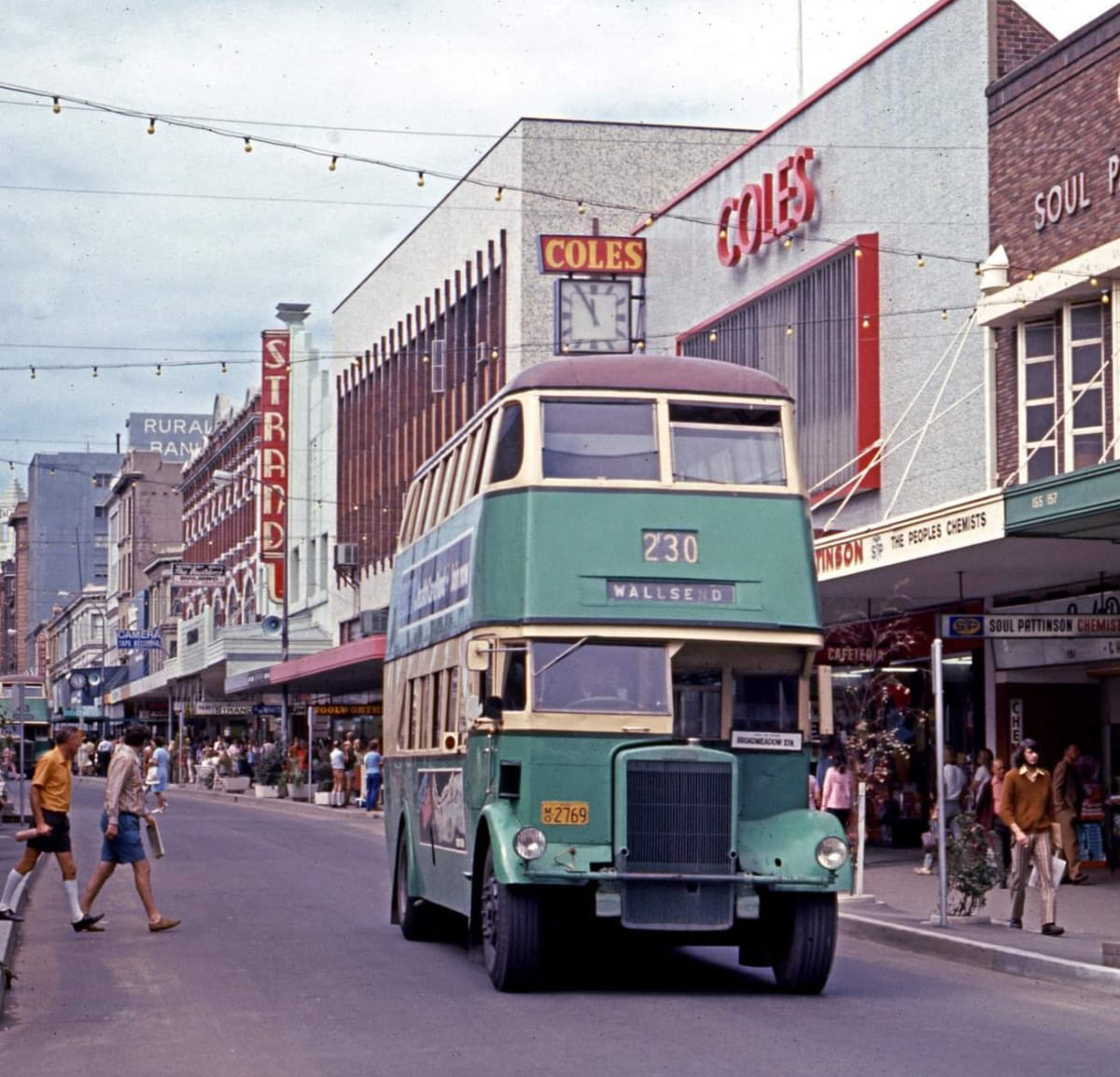 Vintage turquoise double-decker bus on urban street with shopfronts and pedestrians