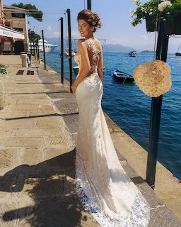 Woman in white lace wedding gown posing on a seaside promenade with blue water and buildings behind