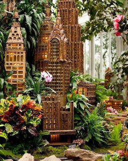Miniature wooden skyscraper models surrounded by tropical plants and orchids in a greenhouse setting