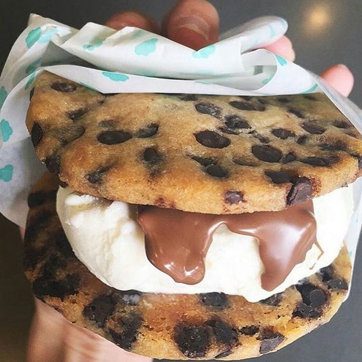 Chocolate chip cookie ice cream sandwich held in hand with white ribbon and melting cream