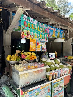 Rustic outdoor fruit stand in Thailand with tropical fruits, coconuts, and colorful signs under wooden shelter
