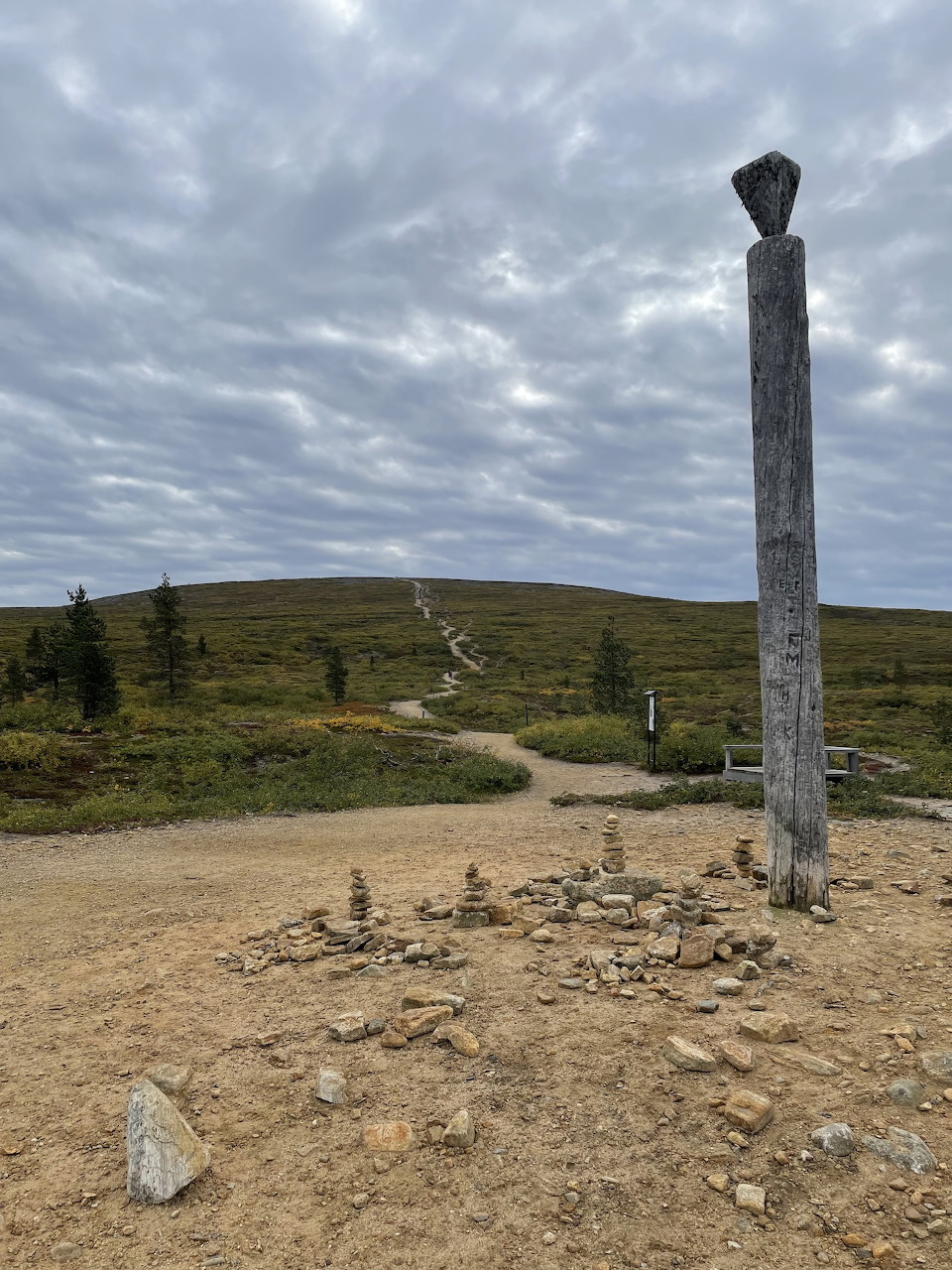 Wooden trail marker post with stone cairns at Kiilopää fell, Lapland, winding path in background