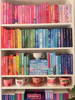 Books organized by color on white shelves with pink mugs below