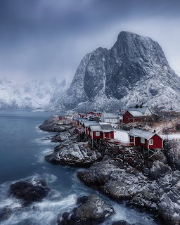 Red wooden cabins on rocky shore beneath snow-covered mountains and overcast skies in Lofoten Norway
