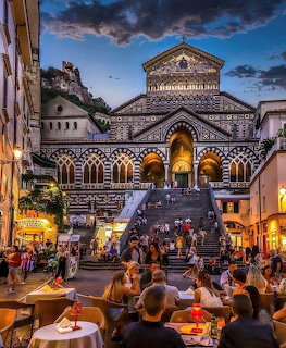 Amalfi Cathedral lit up at dusk with people on the steps and outdoor cafe diners in the foreground