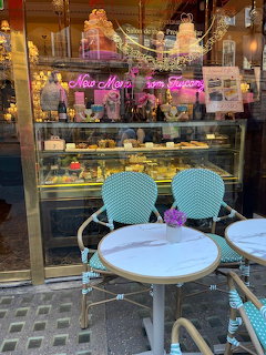 Pink cafe storefront with pastry display, neon sign, and turquoise bistro chairs at marble tables