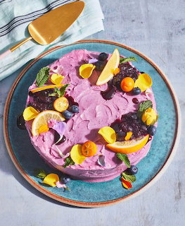 Purple frosted cake on a blue plate topped with citrus slices, berries, and flower petals