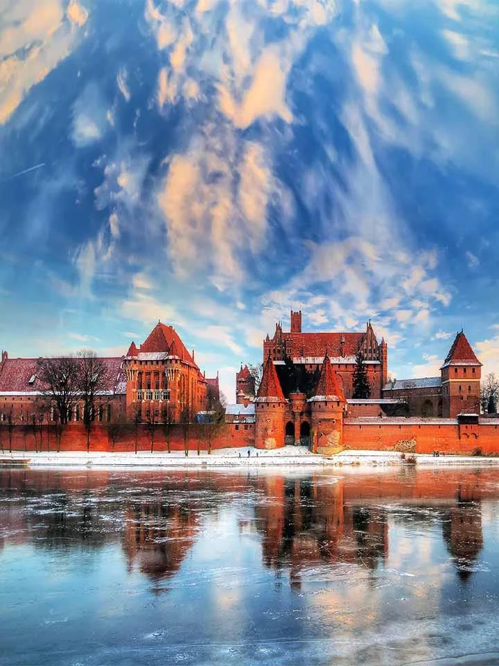 Malbork Castle, a massive red brick medieval fortress in Poland, reflected in an icy river under dramatic clouds.