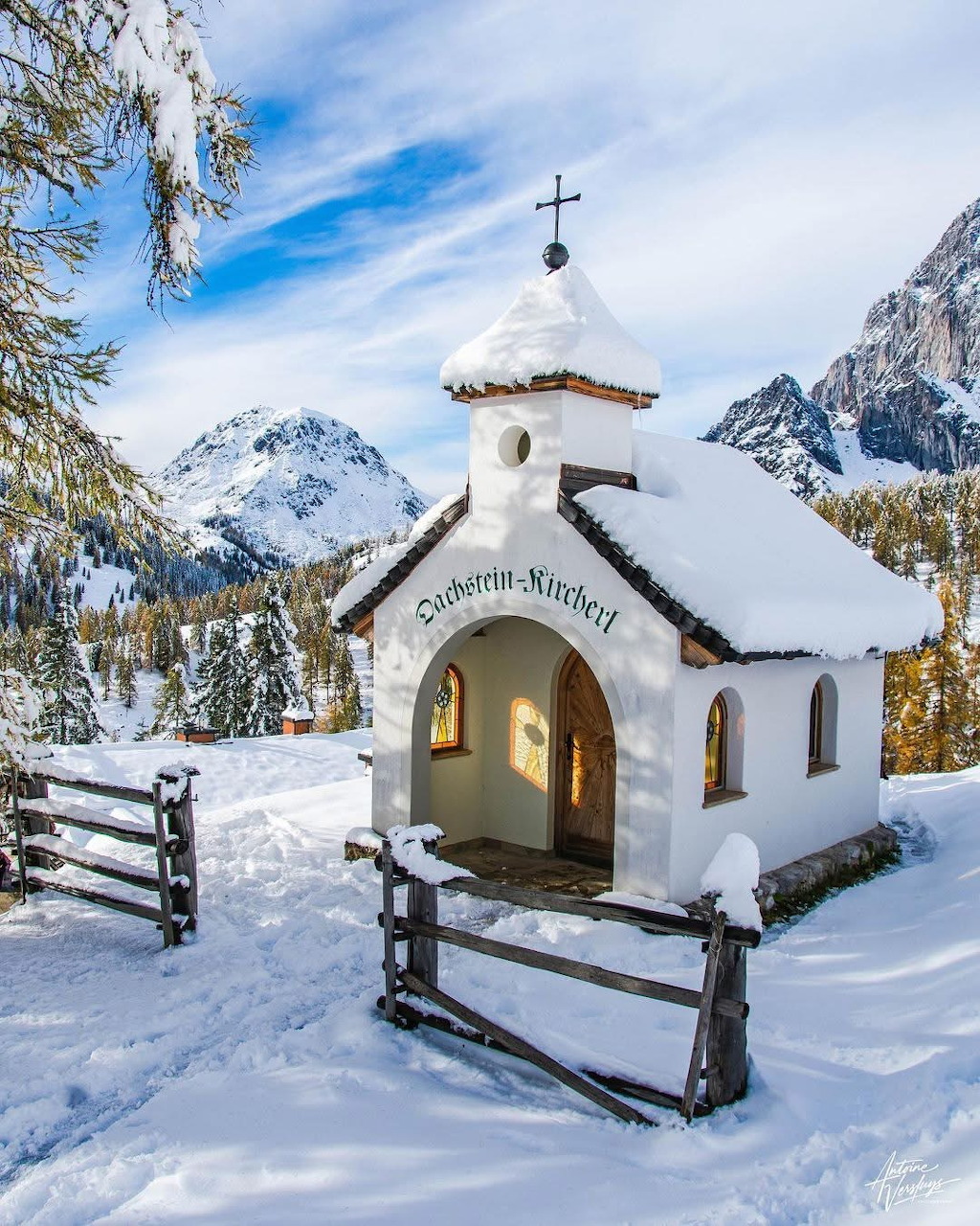 White chapel with cross in snowy mountain landscape surrounded by wooden fence and evergreen trees