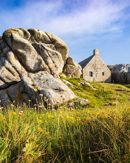 Small stone house wedged between large granite rock formations on a green grassy hillside