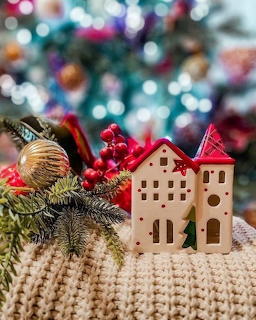 Miniature ceramic house with Christmas decorations including red berries, pine branches, and gold ornament on knitted blanket