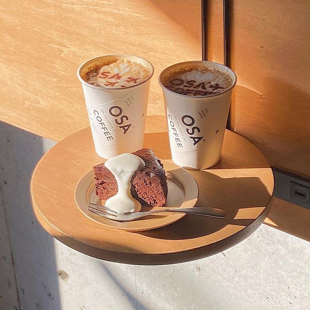 Two paper coffee cups with latte art and a cream-topped dessert on a small round table