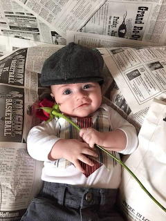 Baby wearing a flat cap lying on newspapers and holding a red rose