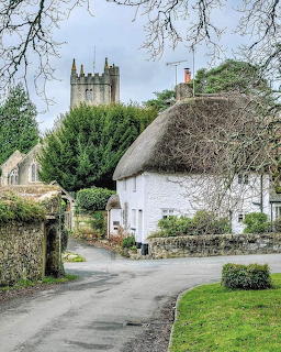 White thatched cottage on village road with stone walls and church tower in Devon