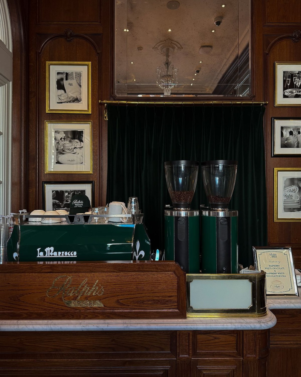 Wooden café counter with green coffee grinders, framed photos on paneled walls, and a crystal chandelier above.