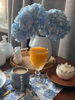 Breakfast table with blue hydrangeas in white vase, orange juice glass, coffee cup, and ceramic decor