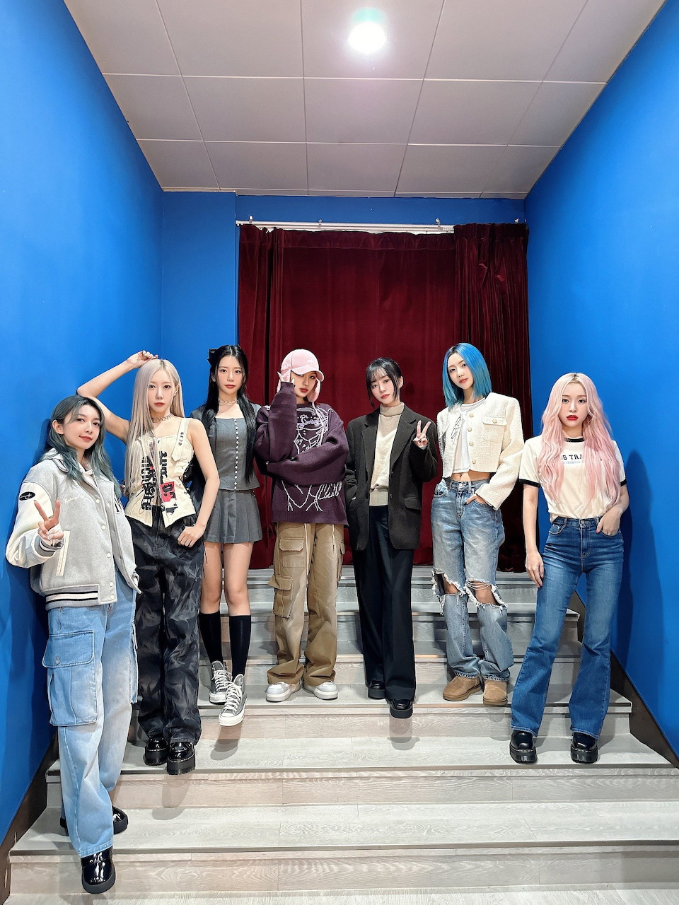 Seven members of K-pop group Dreamcatcher posing together on stairs with a red curtain backdrop