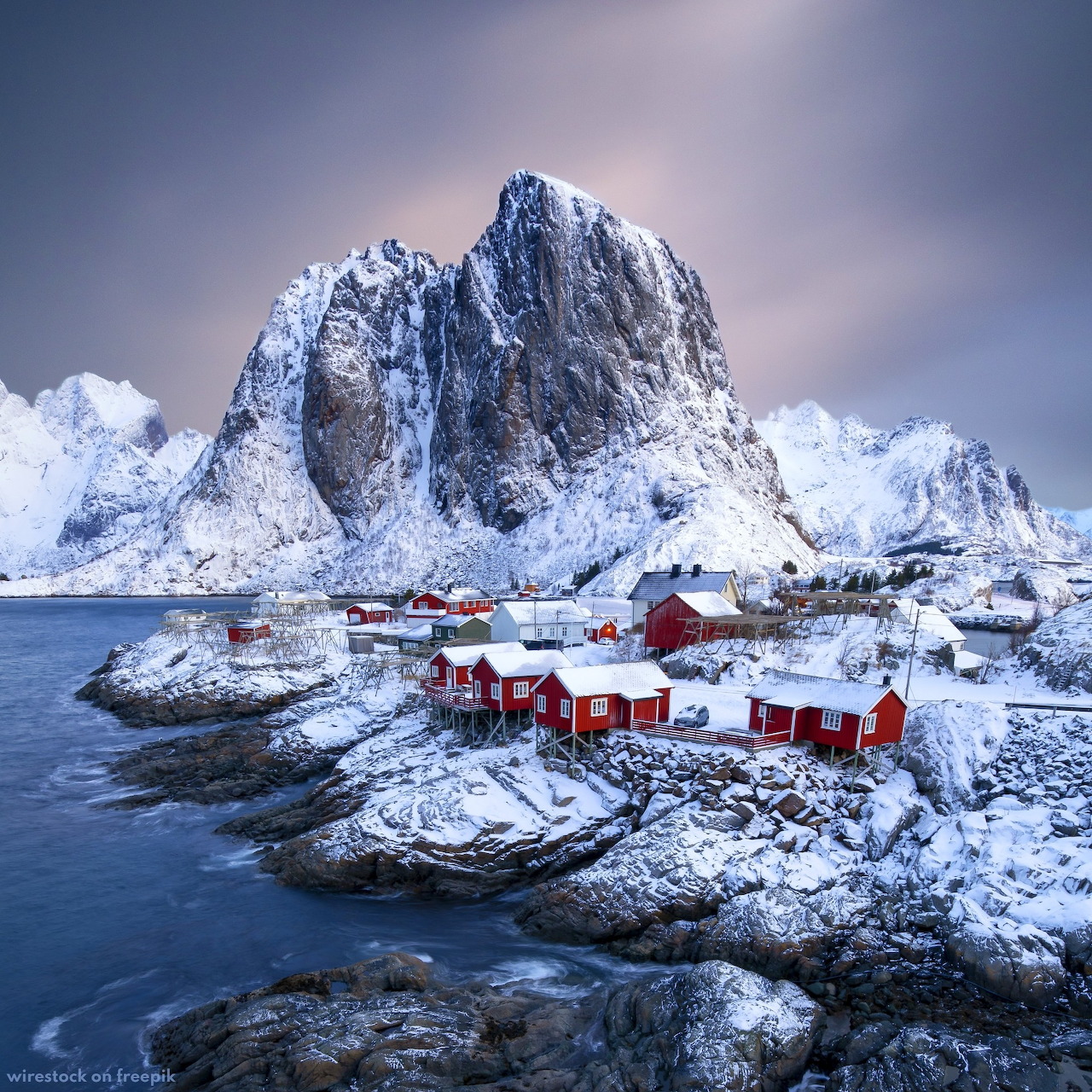 Snow-covered mountain peak behind red fishing cabins on rocky coastal islands in Lofoten, Norway