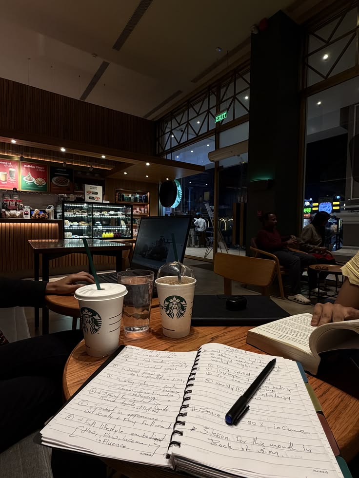 Open notebook and pen beside two Starbucks drinks on a table inside a dimly lit café