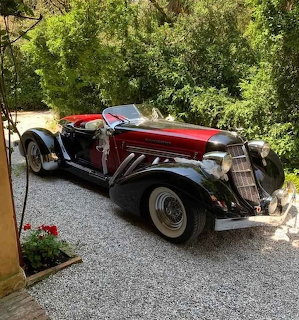 Burgundy and silver vintage Auburn Boattail Speedster convertible on gravel driveway with trees and flowers