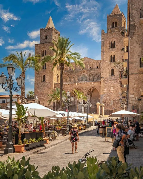 Norman cathedral with twin bell towers in Cefalù, Sicily, surrounded by palm trees and outdoor cafés