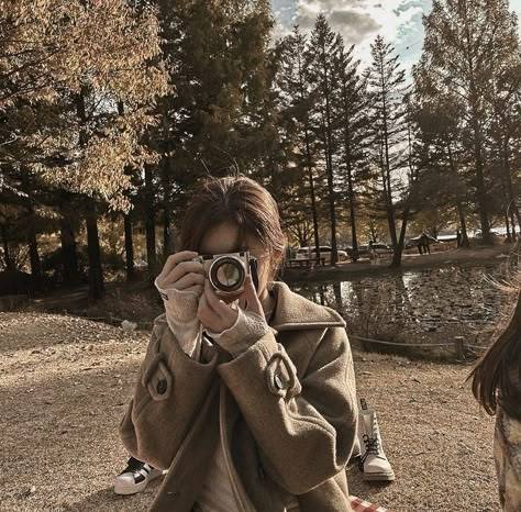 Person in beige coat photographing with a camera in an autumn park with trees and water in background