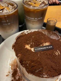 Two iced coffees in plastic cups beside a tiramisu cake dusted with cocoa on a white plate