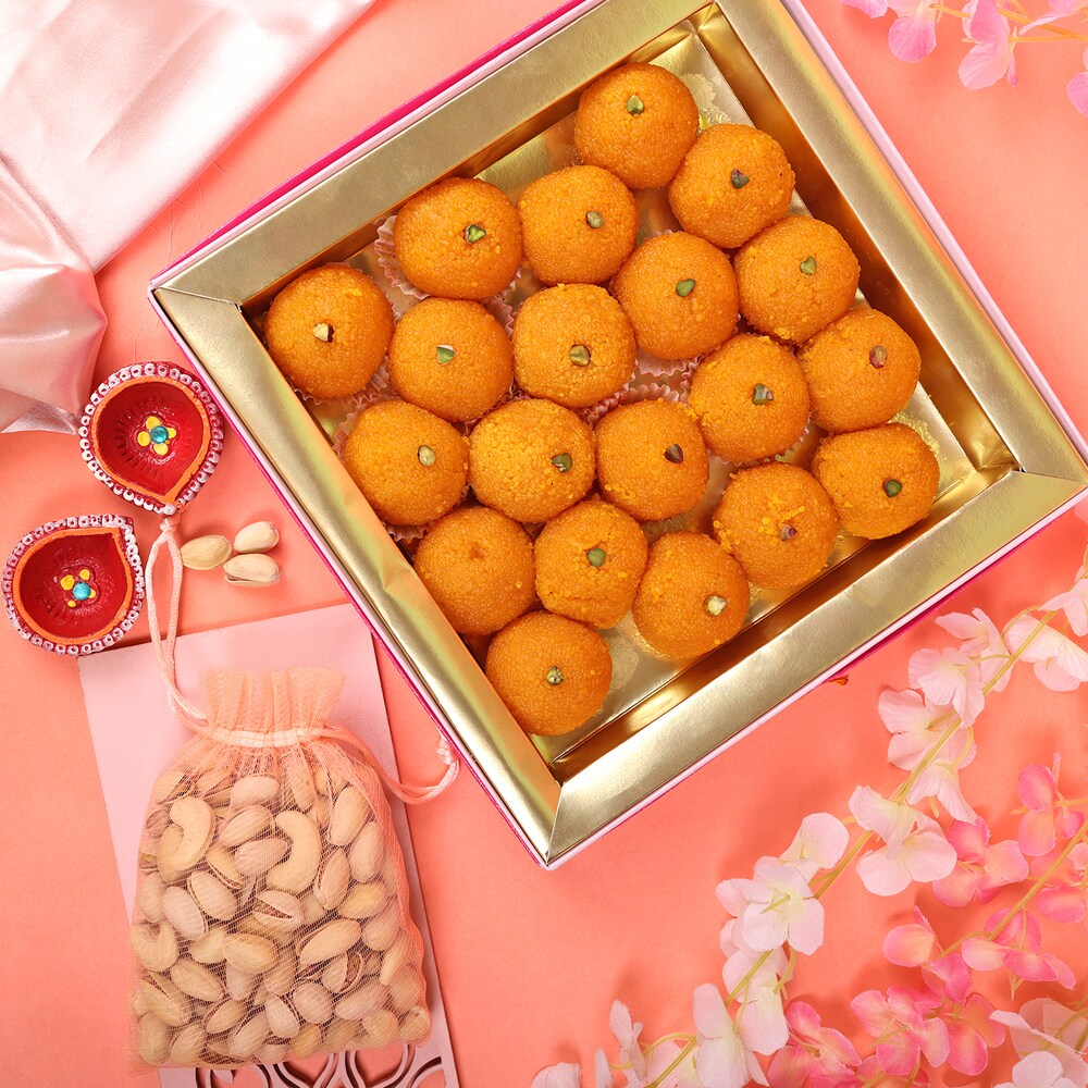 Box of orange ladoo sweets with diyas, cashew bag, and flowers on pink background