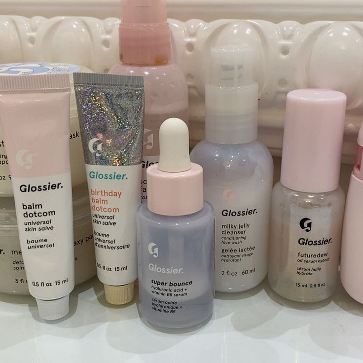 Glossier skincare and beauty products in pastel pink and blue bottles and tubes on a white surface