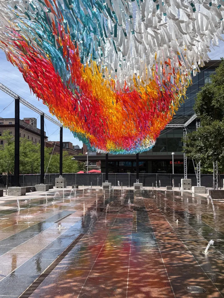 Colorful cascading ribbon sculpture installation above plaza fountain with rainbow gradient from red to blue
