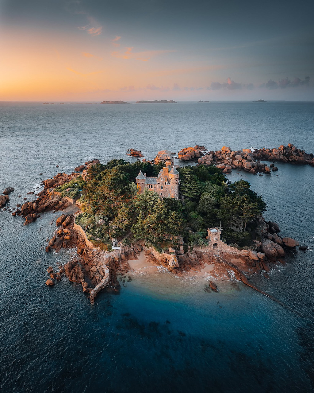 Aerial view of a small rocky island with stone buildings and trees in calm blue water at sunset.