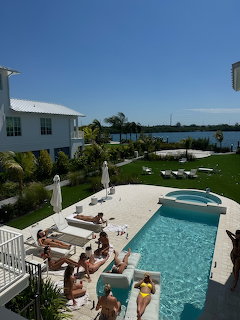 People relaxing by a pool at a white waterfront house with palm trees and a bay in the background