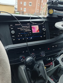 Car dashboard with touchscreen infotainment system showing music playback, gear stick and controls in foreground