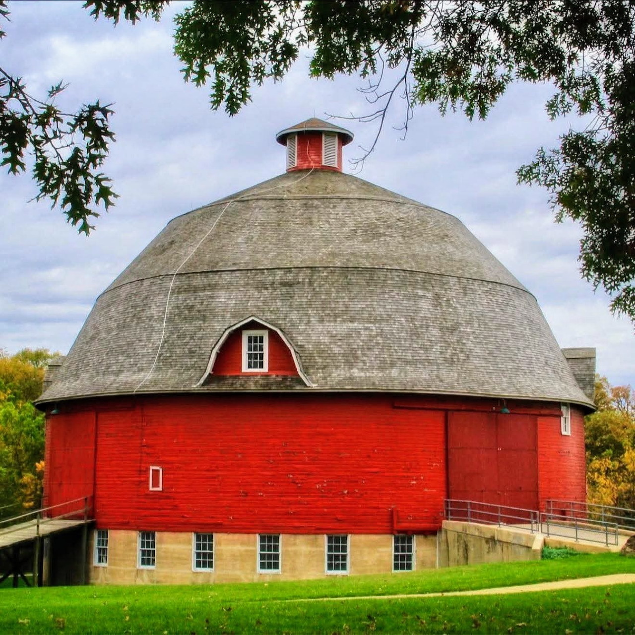Red round barn with domed roof and white cupola on green lawn framed by tree branches