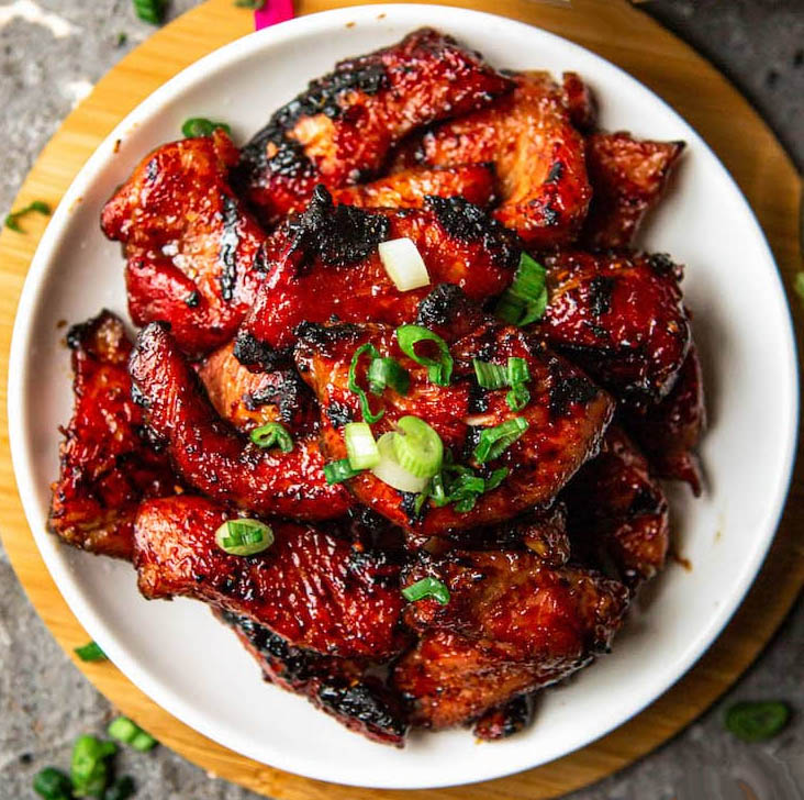 White plate of chared BBQ pork garnished with green onions on a round wooden board