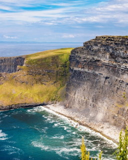 Tall coastal cliffs with layered rock face overlooking ocean waves and green clifftop under blue sky