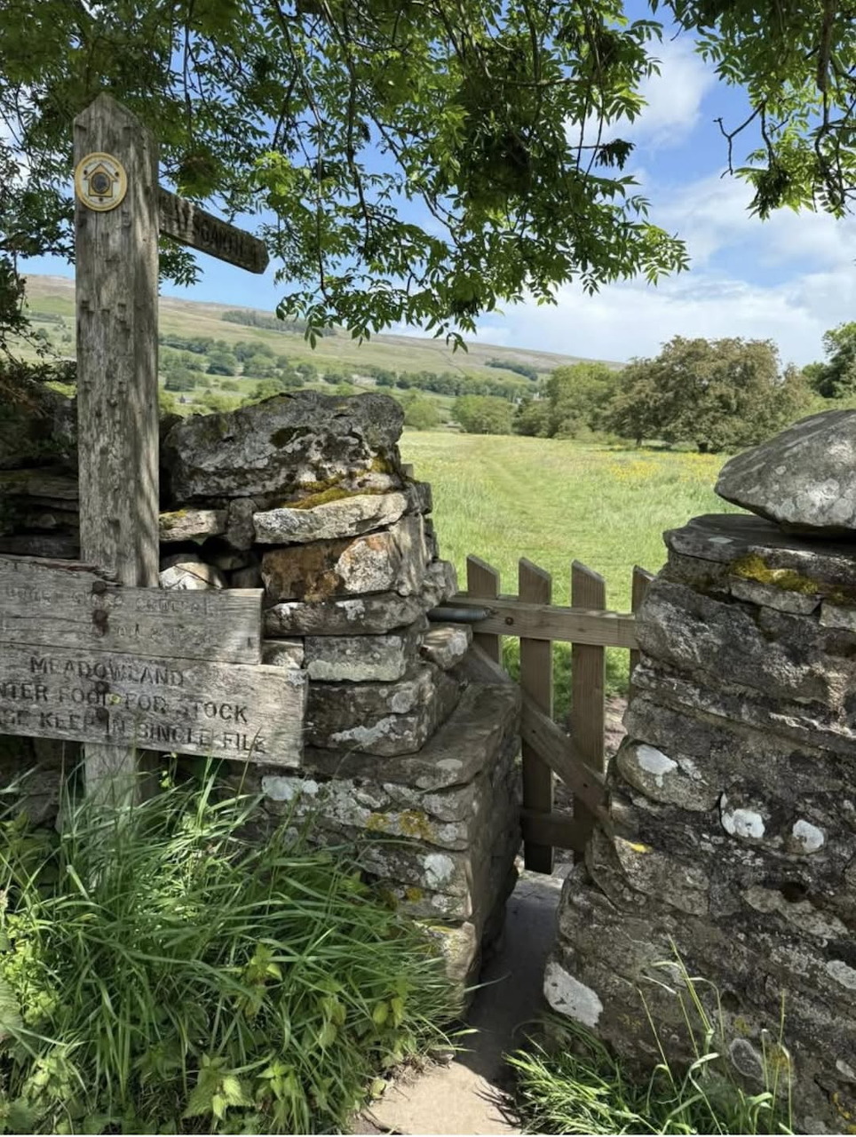 Rustic wooden gate in a dry stone wall with green rolling hills and fields in the background.