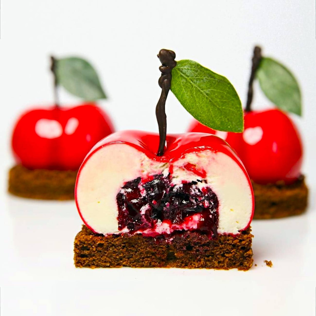 Red cherry-shaped mousse desserts with green leaves on chocolate brownie base, one sliced to show layers