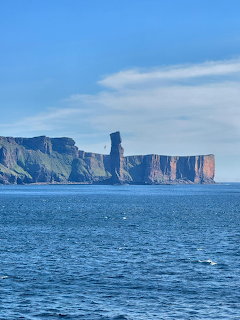 Tall sea stack rock formation rising from ocean with cliffs in background under blue sky