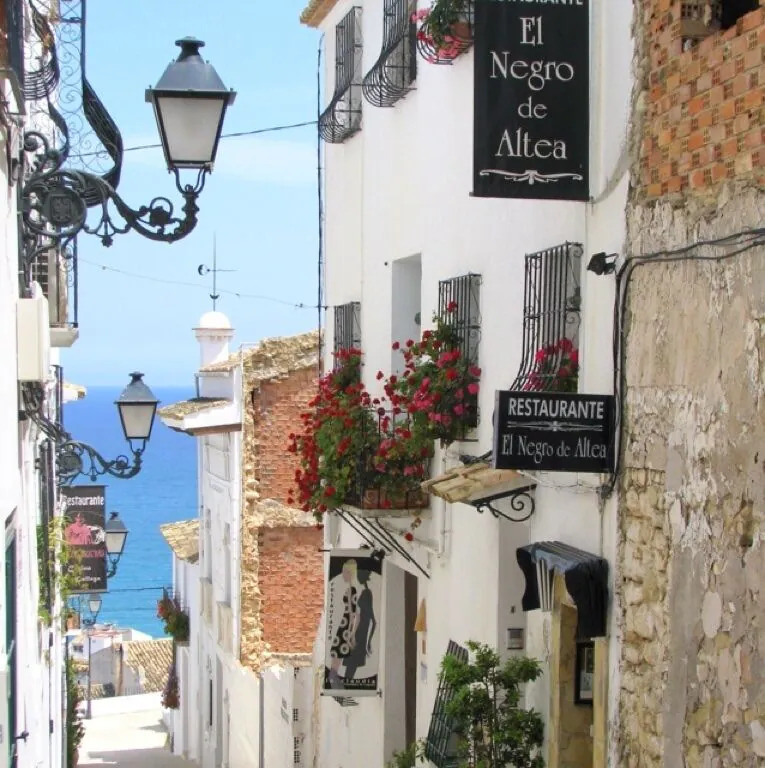Whitewashed street in Altea, Spain with lanterns, flower boxes, and blue sea in the distance