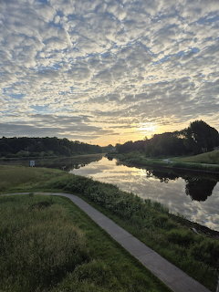 Path beside a calm river at sunset under a cloud-filled sky with grassy banks and trees