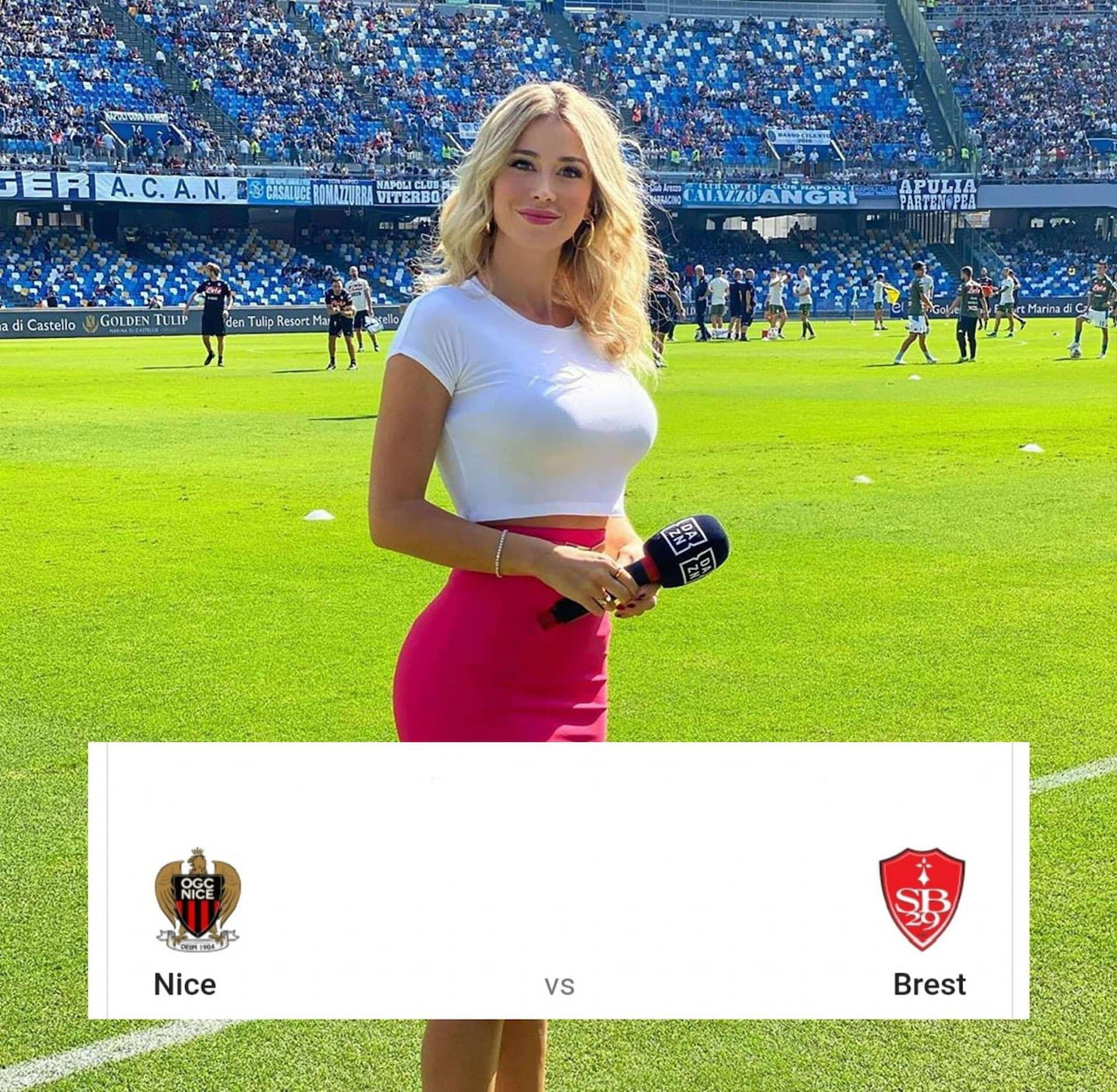 Blonde woman in white top and red skirt posing on soccer field with stadium stands in background