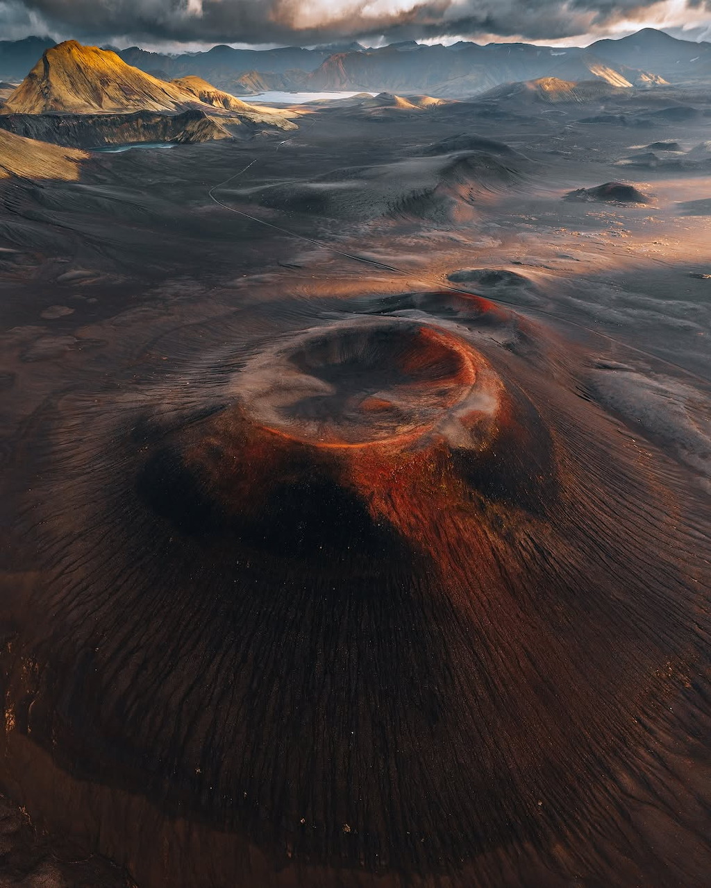Aerial view of a red volcanic crater with dark lava fields and mountains in the background