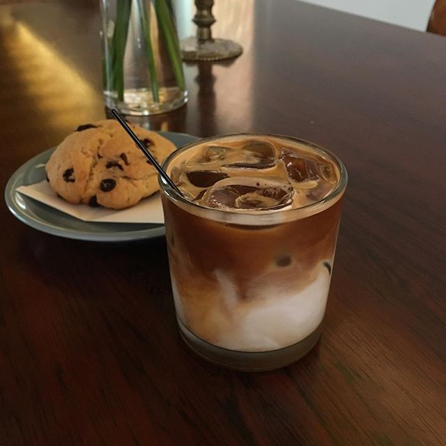 Layered iced latte in a glass on a wooden table with a chocolate chip cookie on a plate behind it.