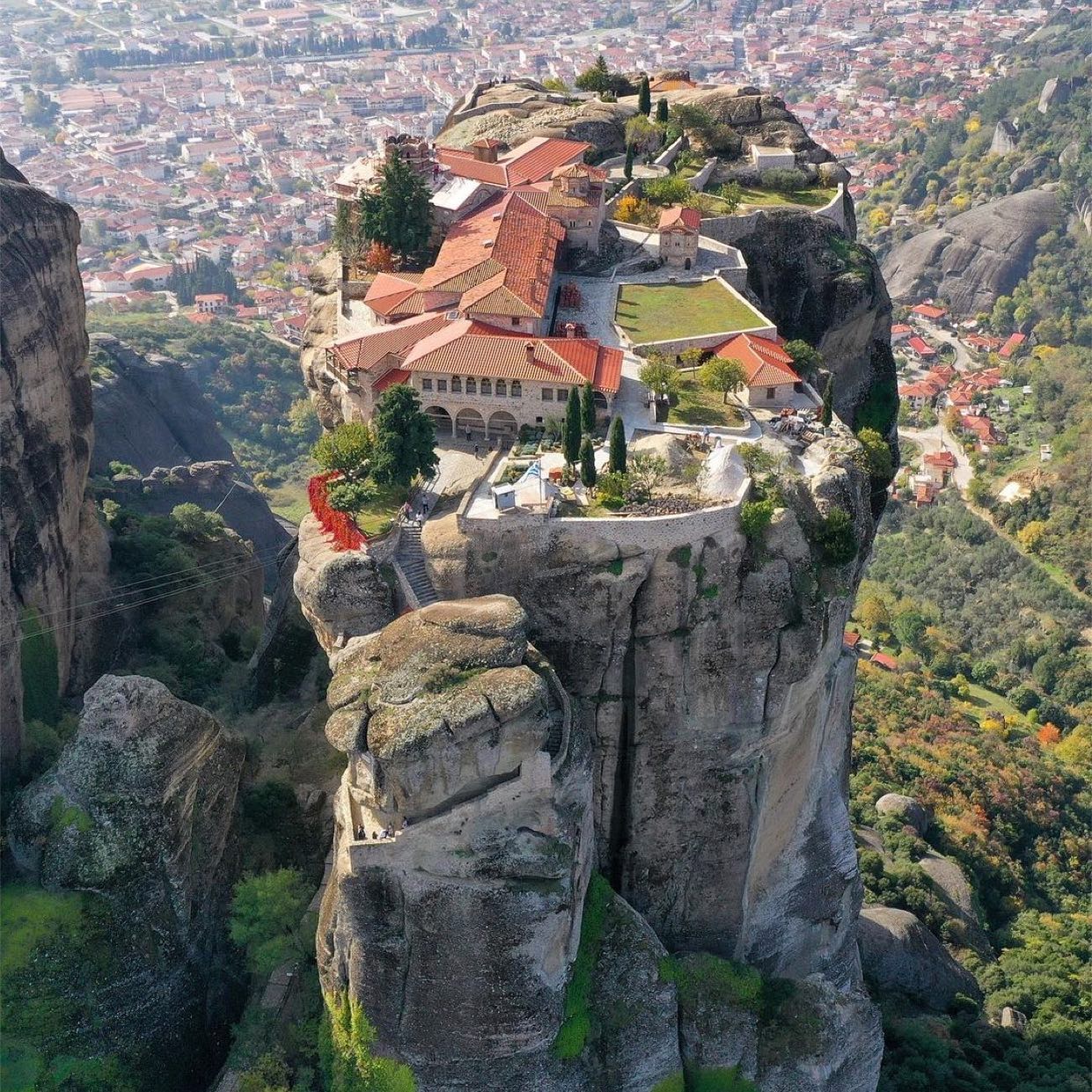 Monastery with red roofs built on top of tall natural rock formations above a green valley and distant town