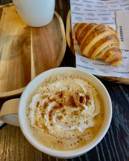 Cup of foamy cappuccino beside a croissant on paper on a wooden table