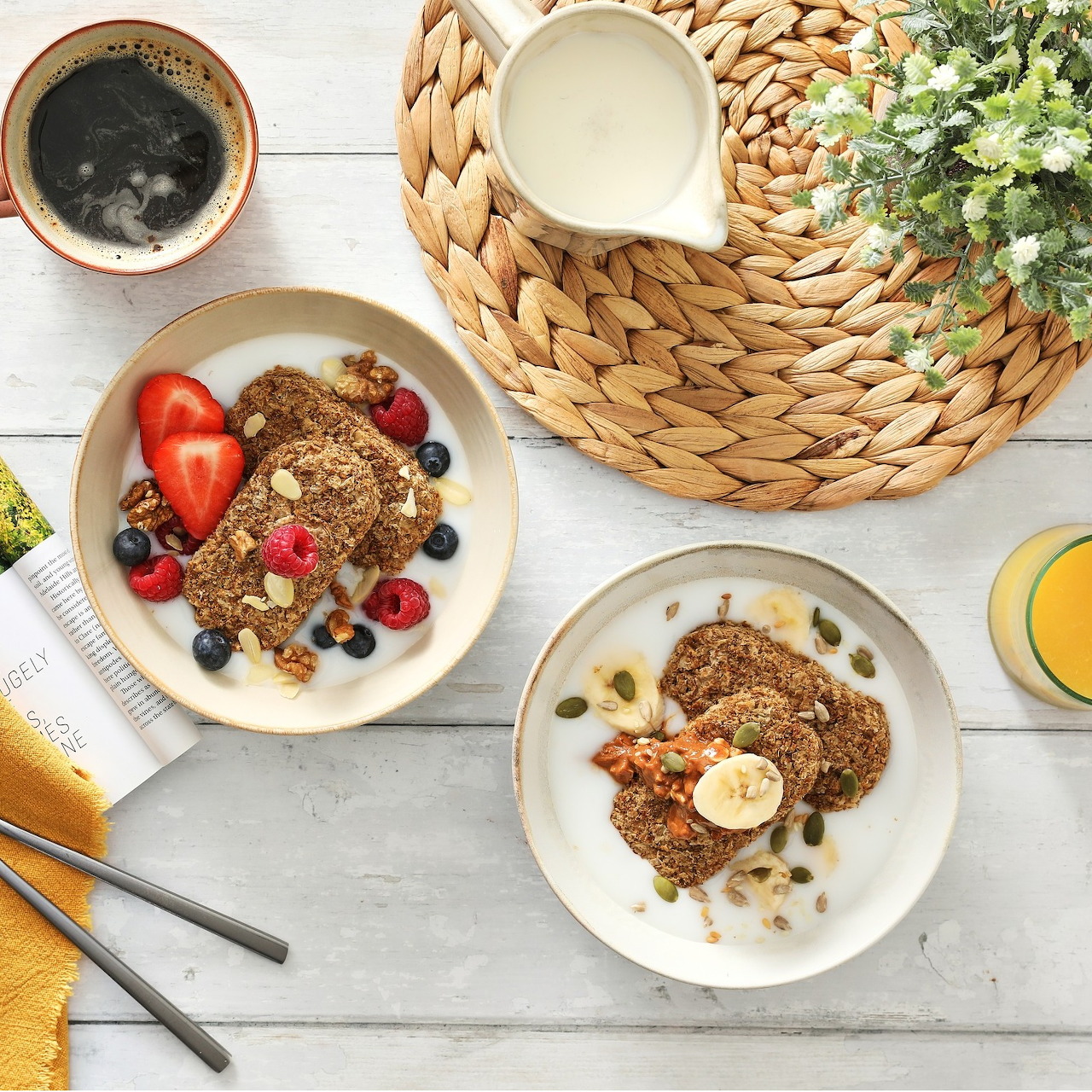 Two breakfast bowls with granola and fresh fruit on a white wooden table with coffee and orange juice