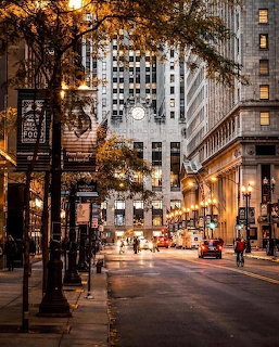 Chicago city street at dusk with lit buildings, autumn trees, and pedestrians under warm streetlights