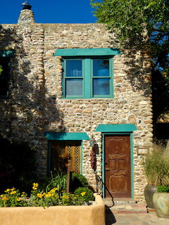 Rustic stone building with teal window trim, carved wooden door, and yellow flowers at the entrance