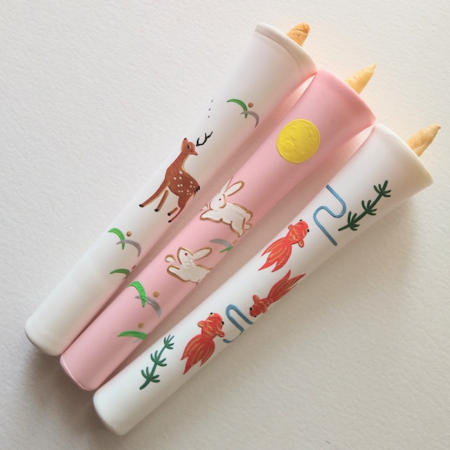 Three decorated pink and white candles with painted rabbits, fish, and a deer on a white surface